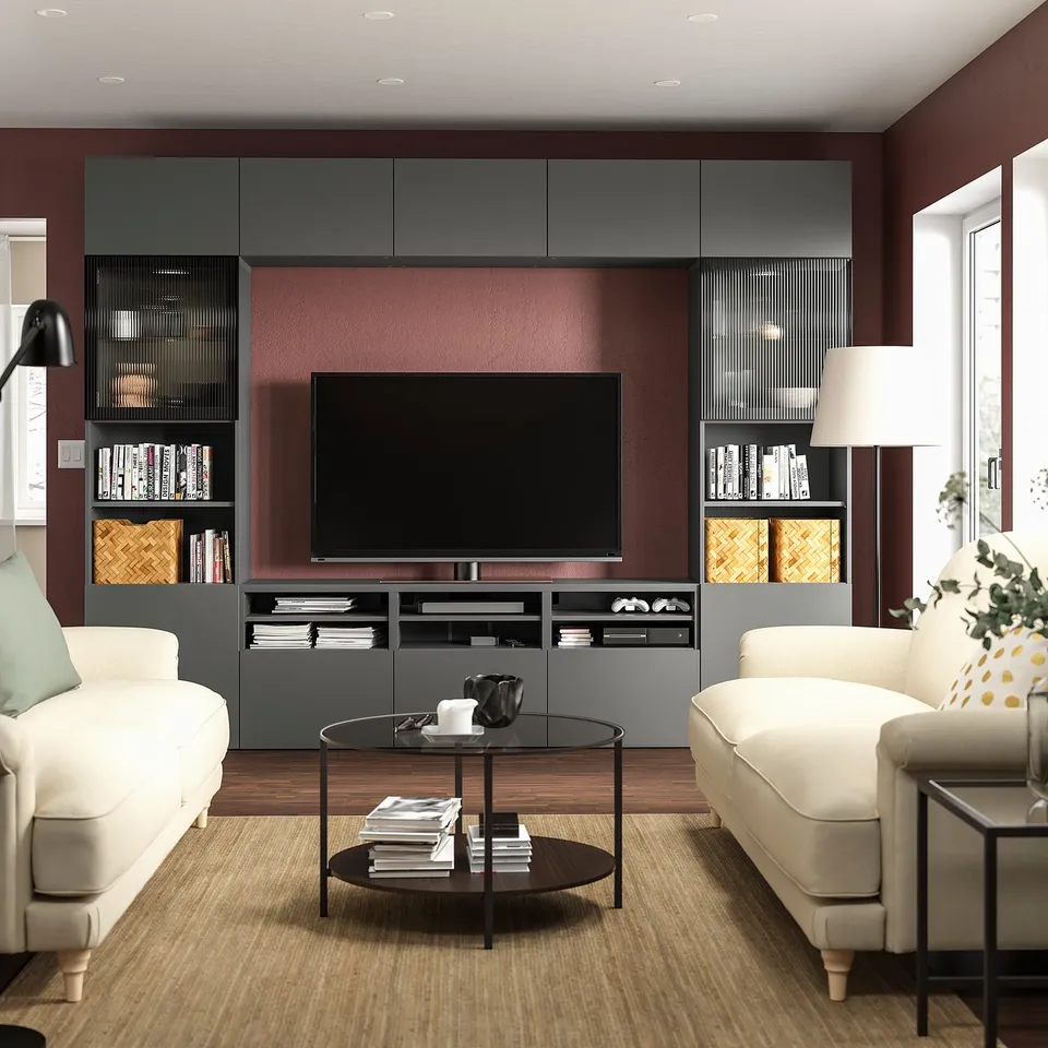 Modern living room with dark red walls, a large flat-screen tv on a grey BESTÅ storage unit, white sofas, a glass coffee table with books and a lamp.