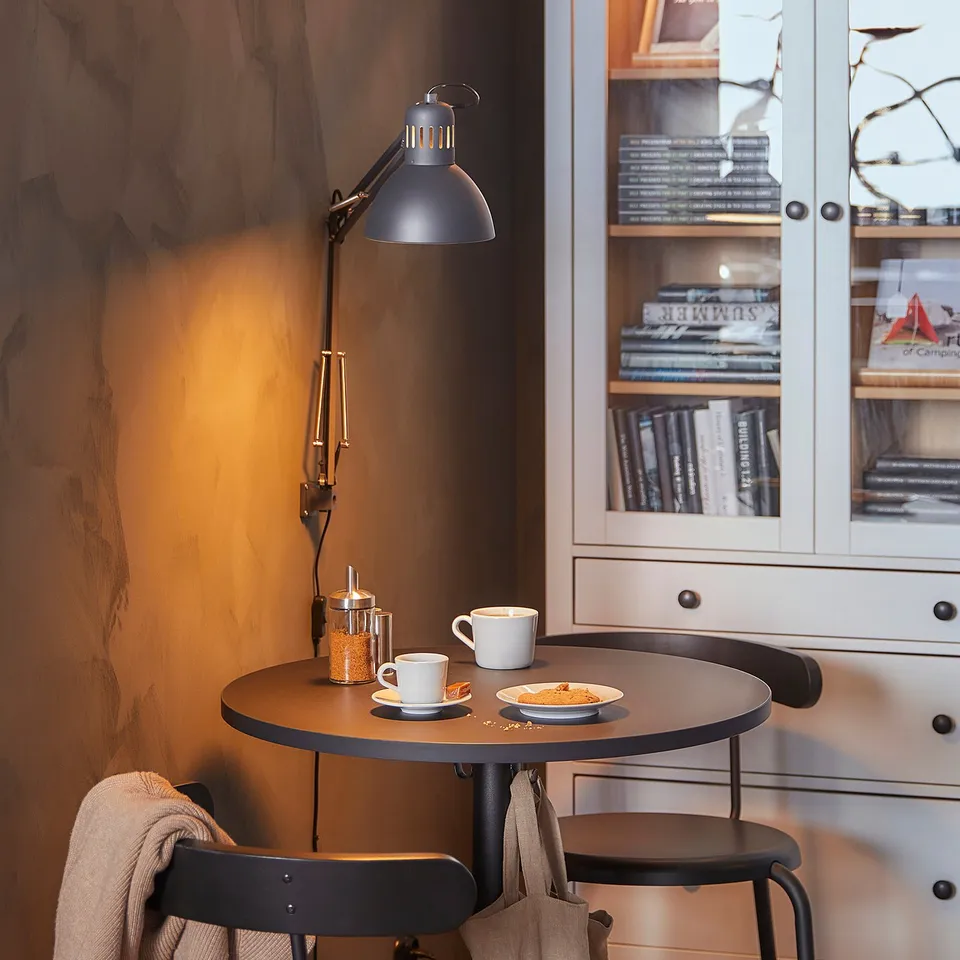 Compact, black, circular dining setup with STENSELE table, wooden top, metal base, hanging lamp, mugs, and chair with draped blanket.
