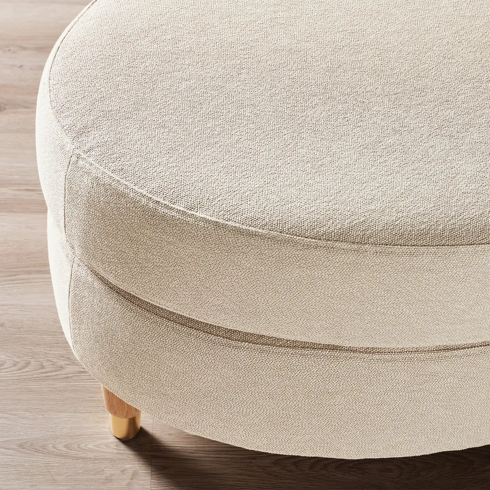 Round, beige upholstered ottoman on wooden floor, quality fabric, sturdy wooden legs.