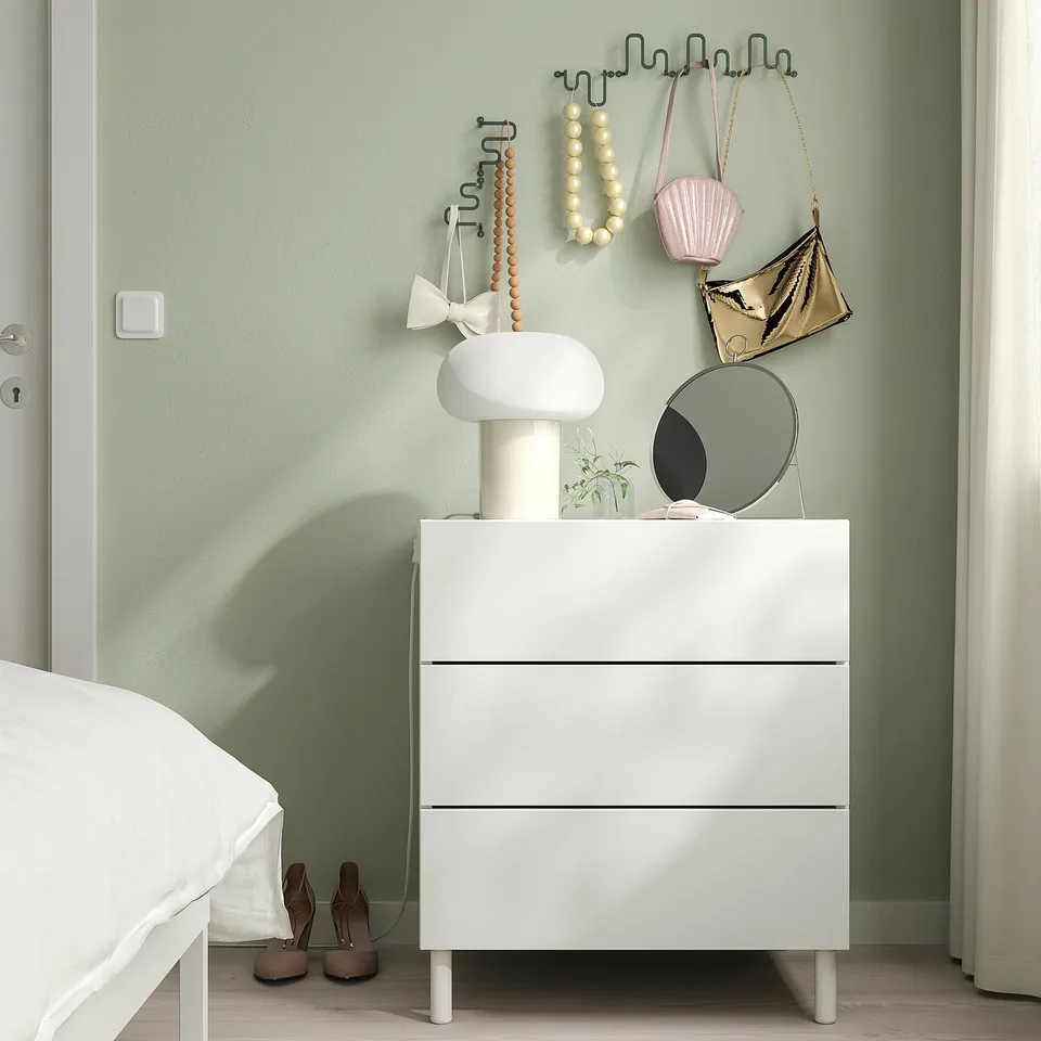 White hook on green wall holding bags & beads, minimalist room with dresser & bed.