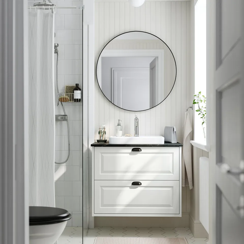 Bright white bathroom: round mirror, sleek white vanity with drawers, black countertop, small sink, and shower with a transparent curtain.