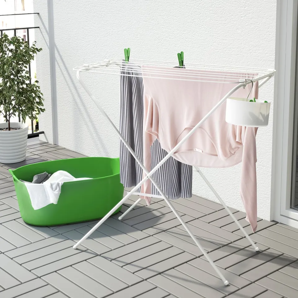 White foldable drying rack with pink clothes on balcony, green bin and plant nearby.