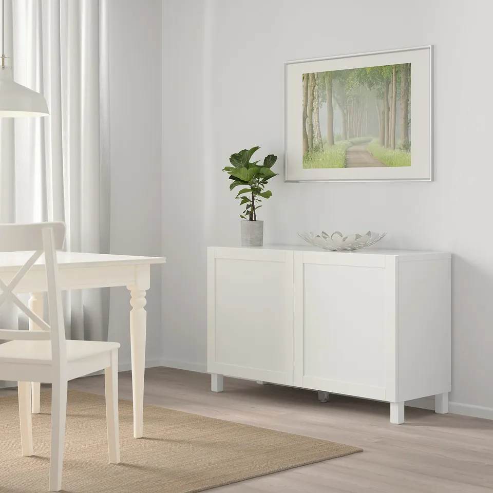 Minimalist room with BESTÅ sideboard, plant, and forest picture. Dining table with white chairs by window.