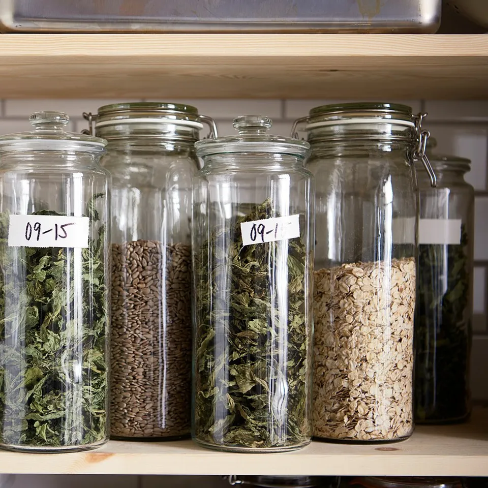 A shelf of clear glass jars with metal clamps stores herbs, beans, and grains. Jars labelled with dates.