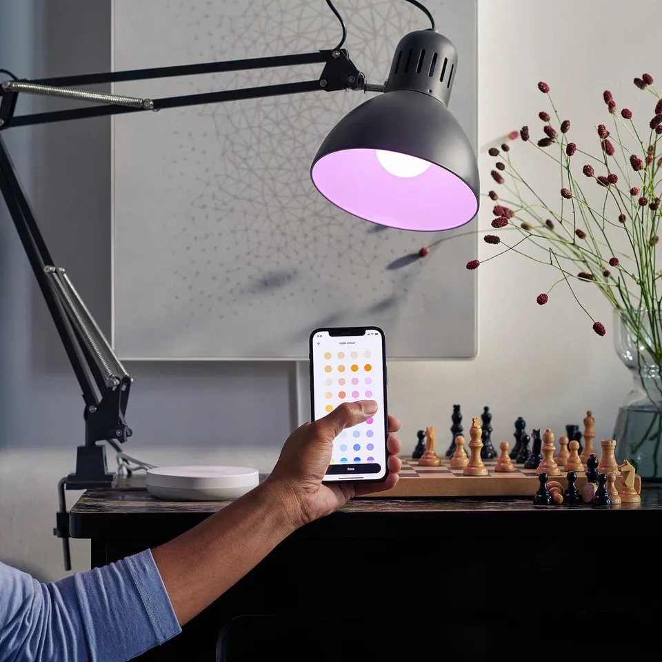 Hand holds phone, changing smart lamps colour via app. Desk has chess set, flowers, and lamp with purple light.
