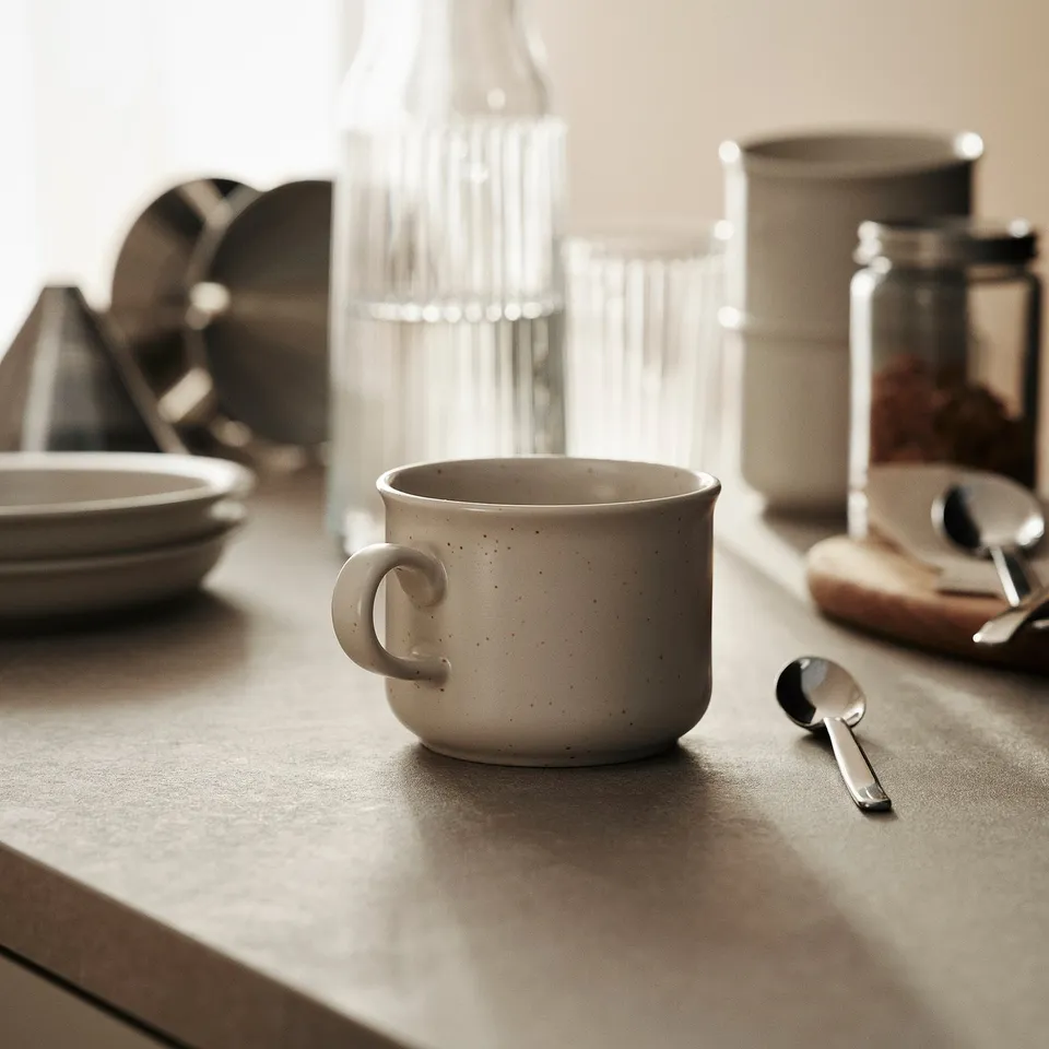 Small, speckled off-white DAGGASTER mug on table with spoon, coffee setup, and plates.