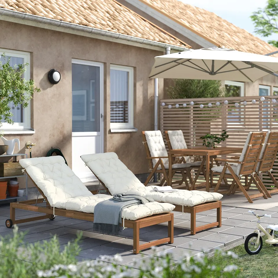 Backyard with NÄMMARÖ tan reclining chairs, wooden table, and beige umbrella. House with tiled roof in distance.