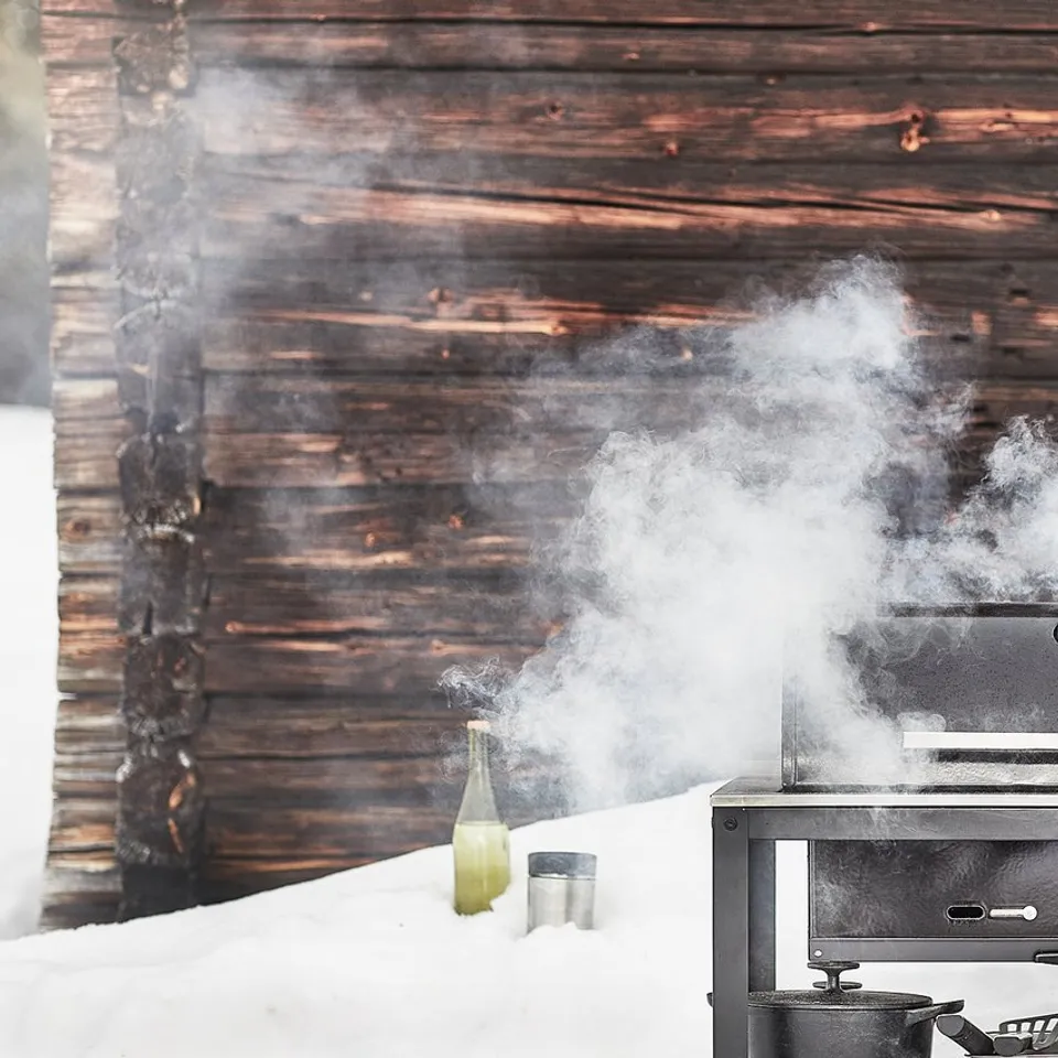 A person grills outdoors in snowy weather using a black GRILLSKÄR grill. Steam rises from the grill, and a wooden cabin is visible in the background.