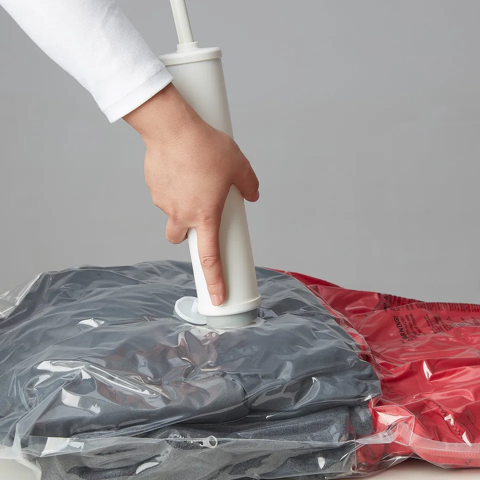 Person uses white manual vacuum pump to remove air from sealed plastic bag. Helps compress clothing for storage.