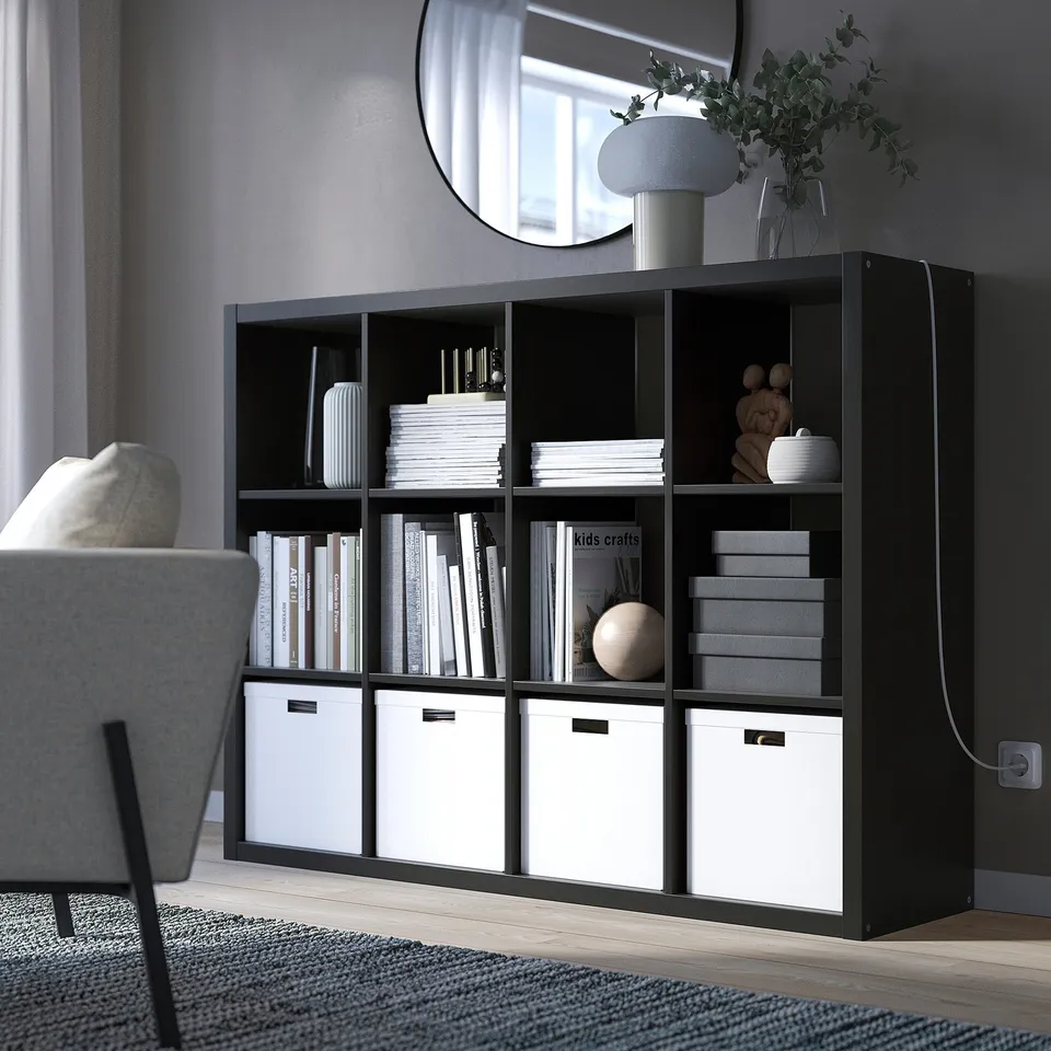 Black KALLAX shelf with books, boxes, decor next to grey chair.