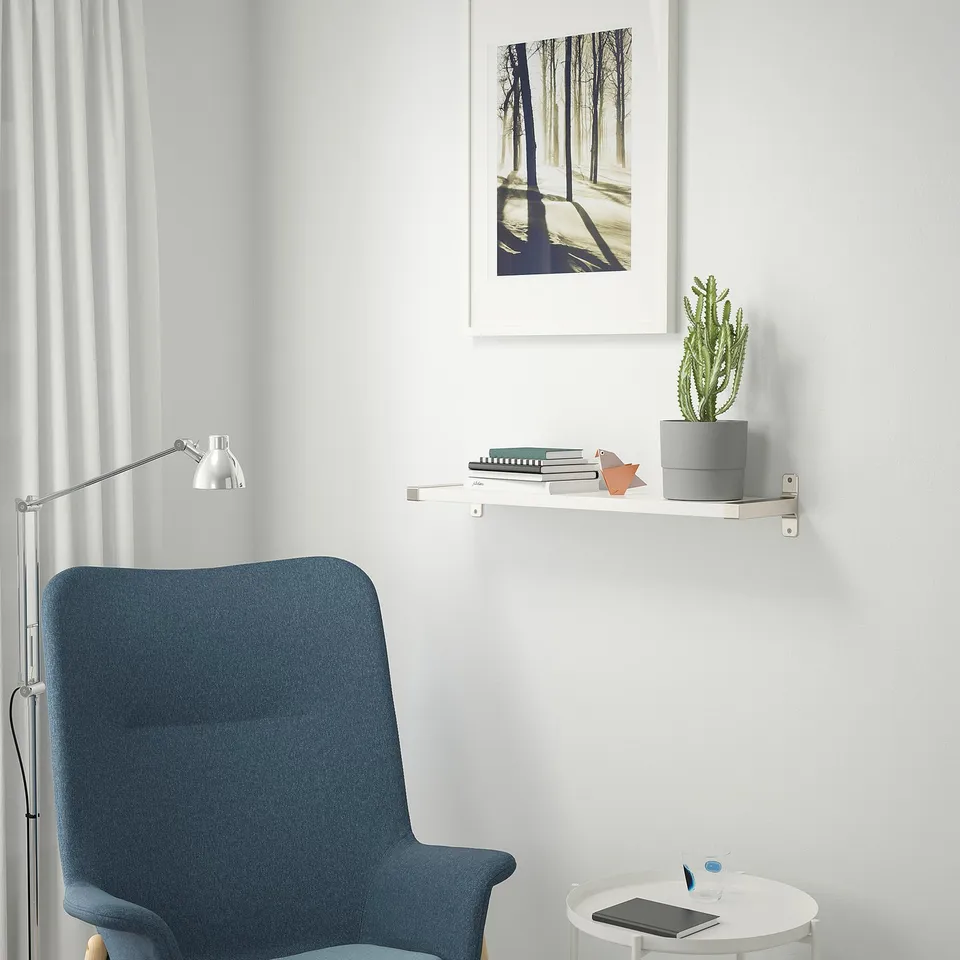 Modern study area with blue chair, wall shelf, books, cactus, art, lamp, and table with tablet and pen.