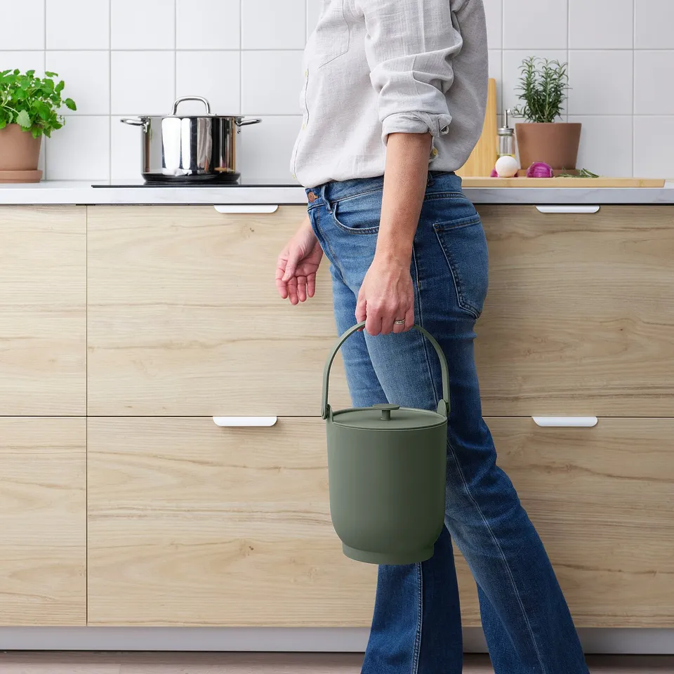 Person holding a green kitchen bin with a handle. The bin is designed to collect food scraps and is easy to clean. It has a lid to avoid odours.