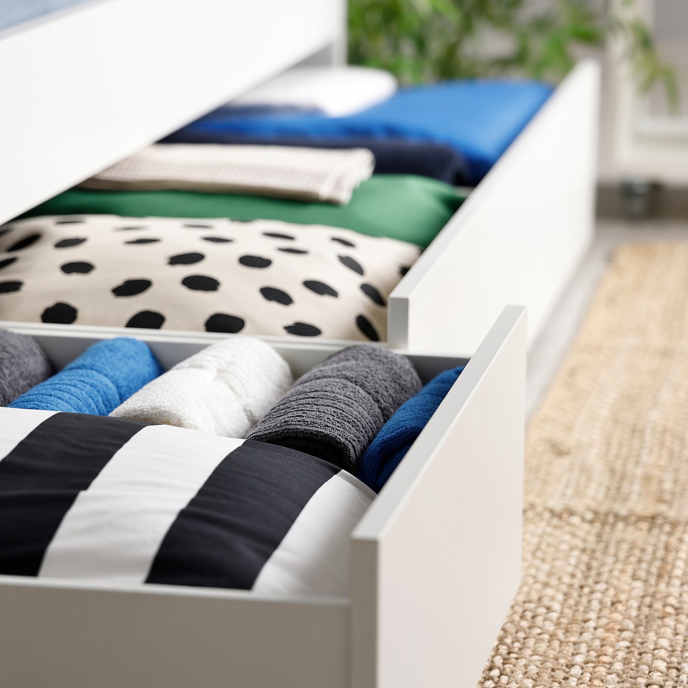 Open drawer reveals neatly organised bed linens: folded blankets, pillows, and sheets in various colours.