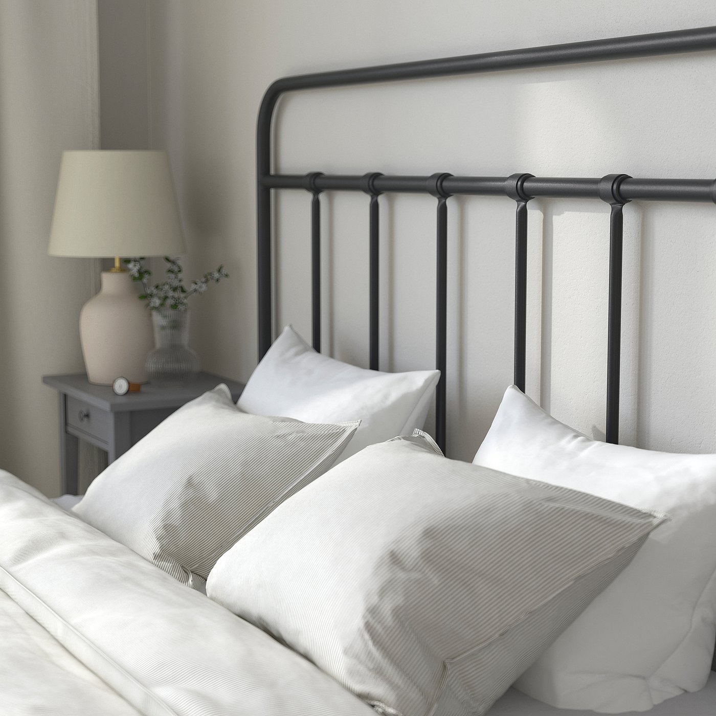 Bedroom scene: white bed with black metal headboard, white pillows, and grey bedside table with lamp and vase.