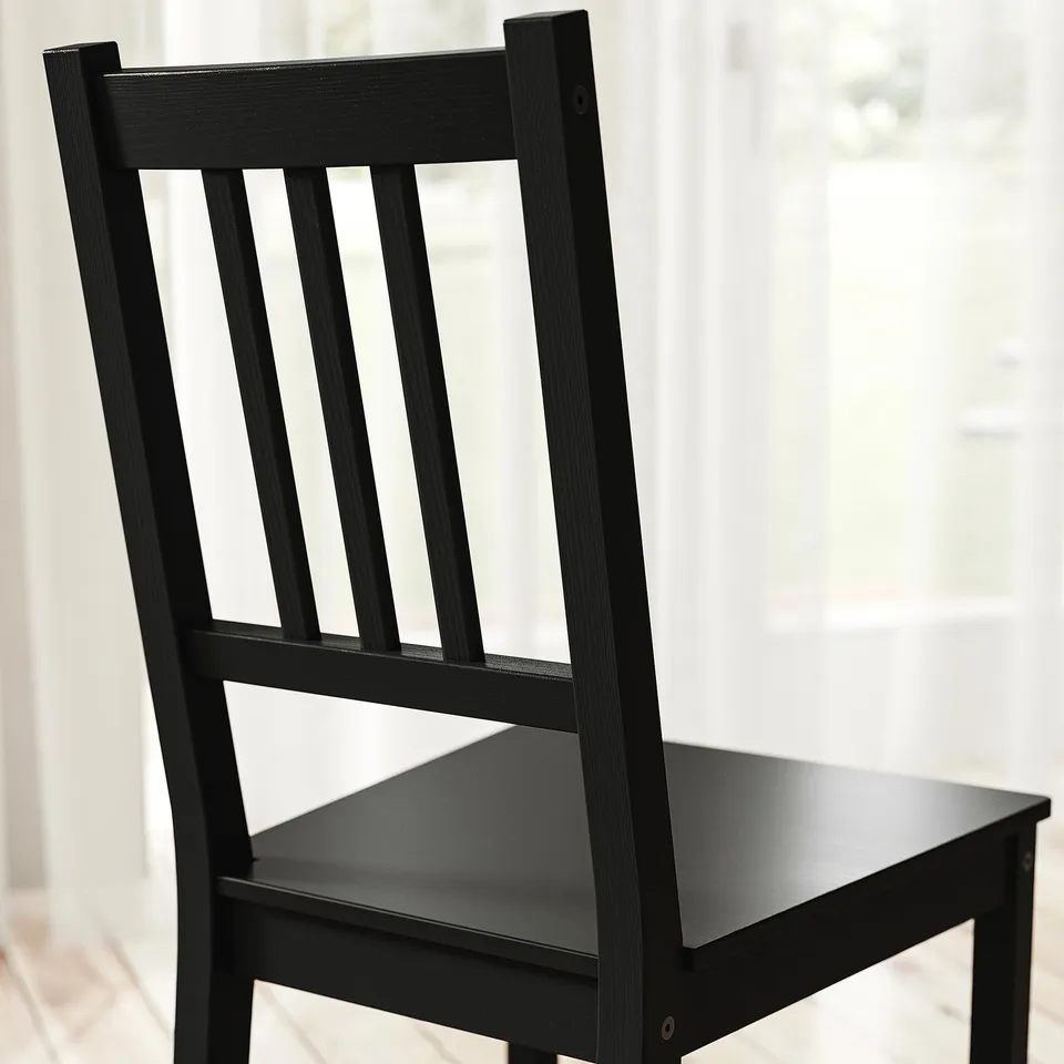 Black wooden chair with vertical slatted back and seat, sturdy and well-finished, standing against a bright window.