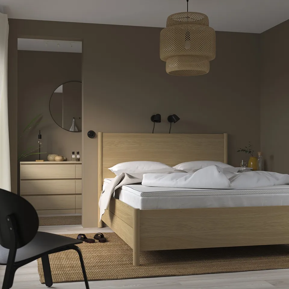 Minimalist bedroom with light wooden bed, white linens, hanging lamp, and black chair. Bed is larger than chair, placed centrally.