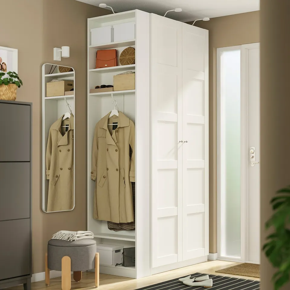 A white PAX wardrobe end-unit beside a door. It has narrow and tall, with garments hanging and accessories on shelves.