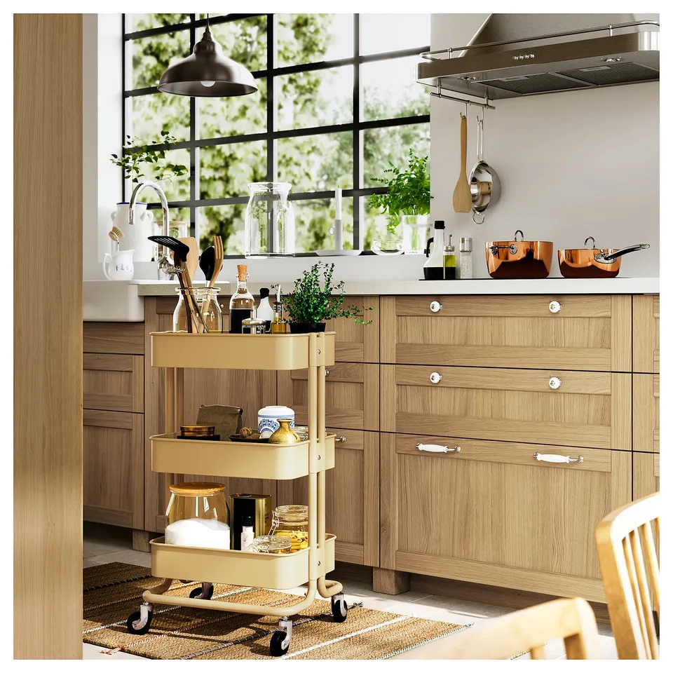 Kitchen with wooden cabinets and drawers, a yellow cart with items, white knobs too. Large window, plants, hanging ladles, bright and cosy.
