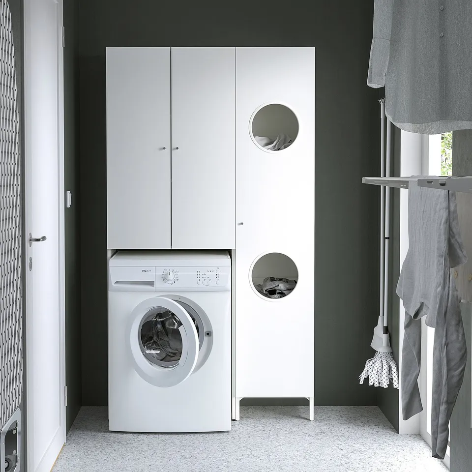 Nycjn laundry setup: white machine under tall cabinet with circular cutouts, dark green walls, white speckled floor.
