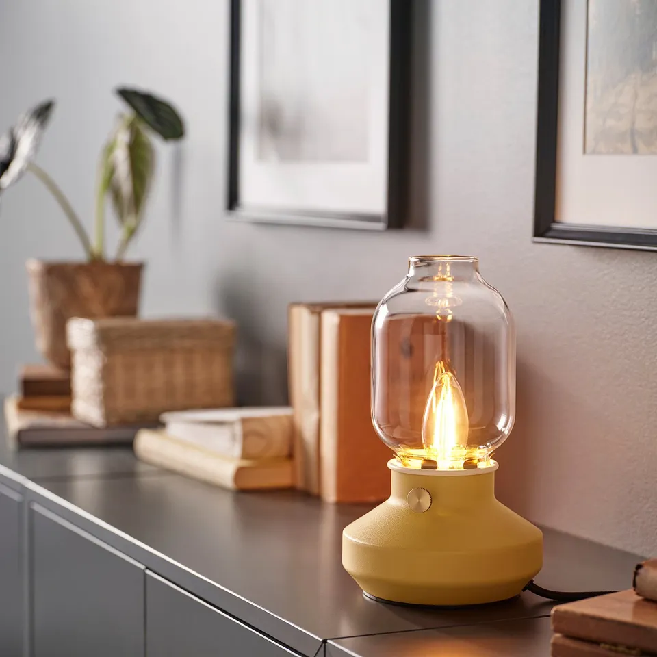 Small yellow TÄRNABY lamp sits on a shelf, glowing warmly with a visible bulb. Its surrounded by books and a potted plant, near framed pictures.