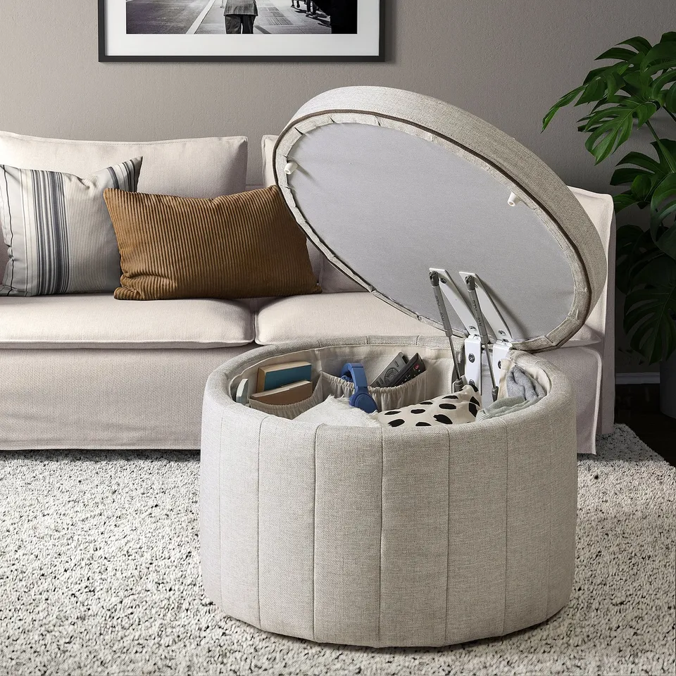 Modern living room with white couch, pillows, and ottoman. The ottoman is open, revealing storage compartments filled with items.