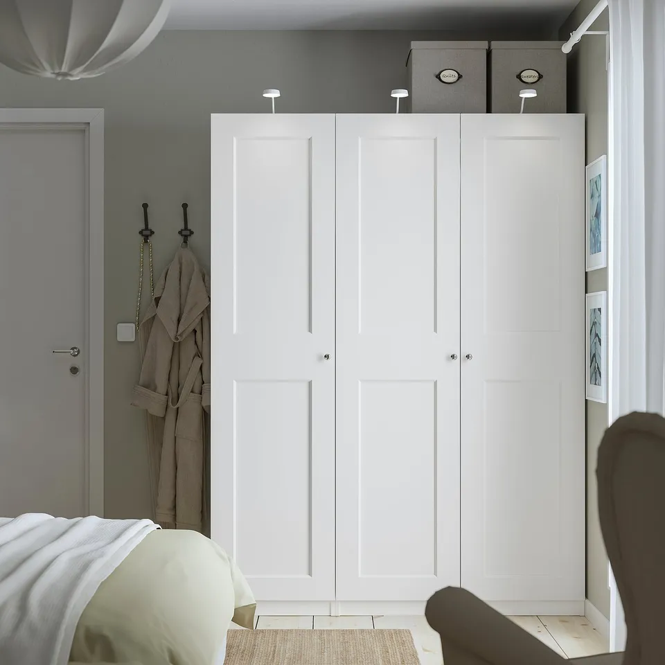 Minimalist bedroom with large white PAX wardrobe, silver handles, two grey boxes, narrow door, bed with white and beige linen.