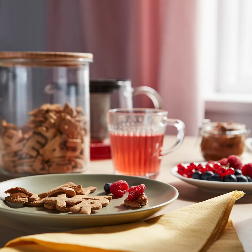 A breakfast table with KAFFEREP letter biscuits on a green plate, fresh berries, and a glass mug of red tea, set on a yellow cloth.