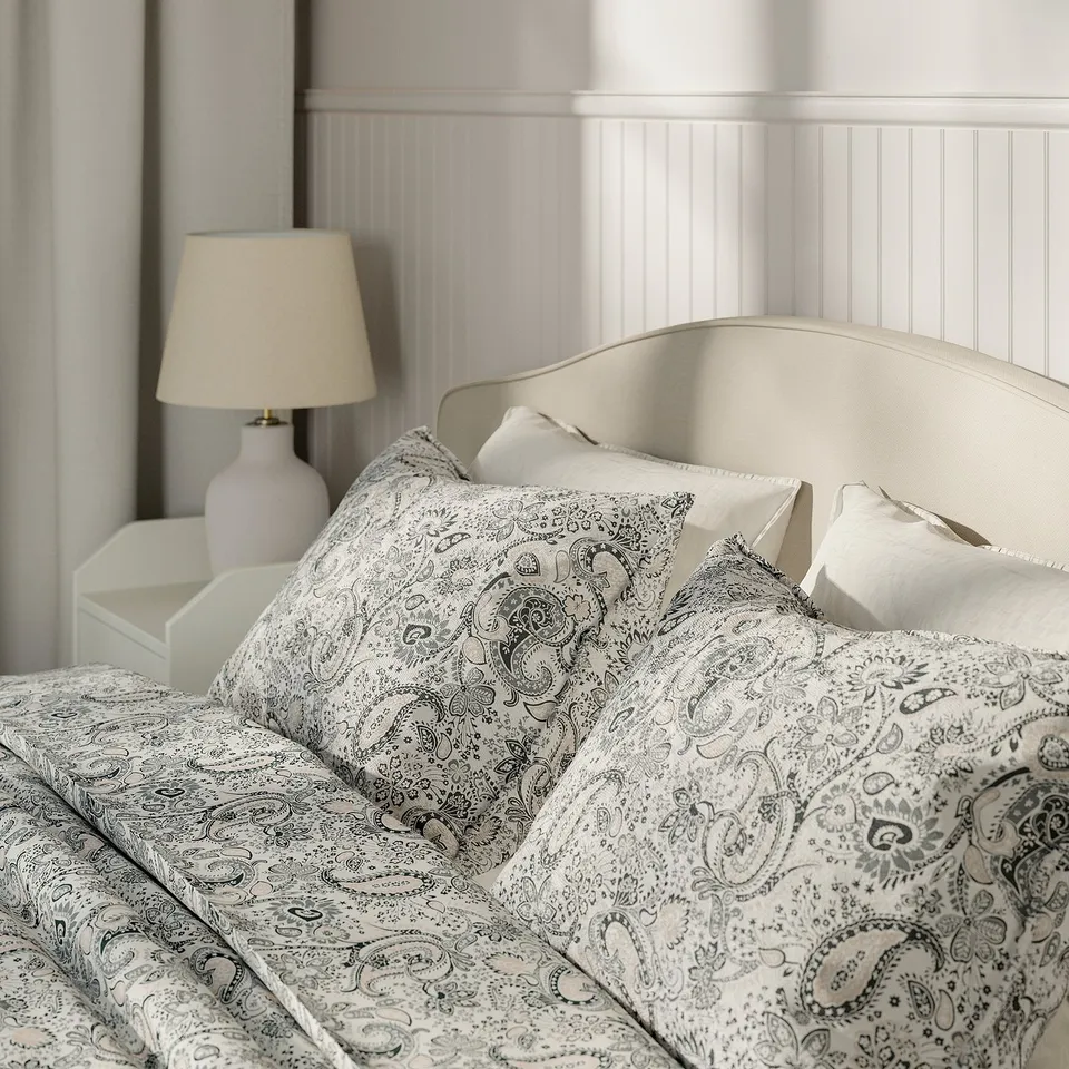 Bedroom scene with a beige upholstered bed, patterned pillows, and a bedside table with a lamp.