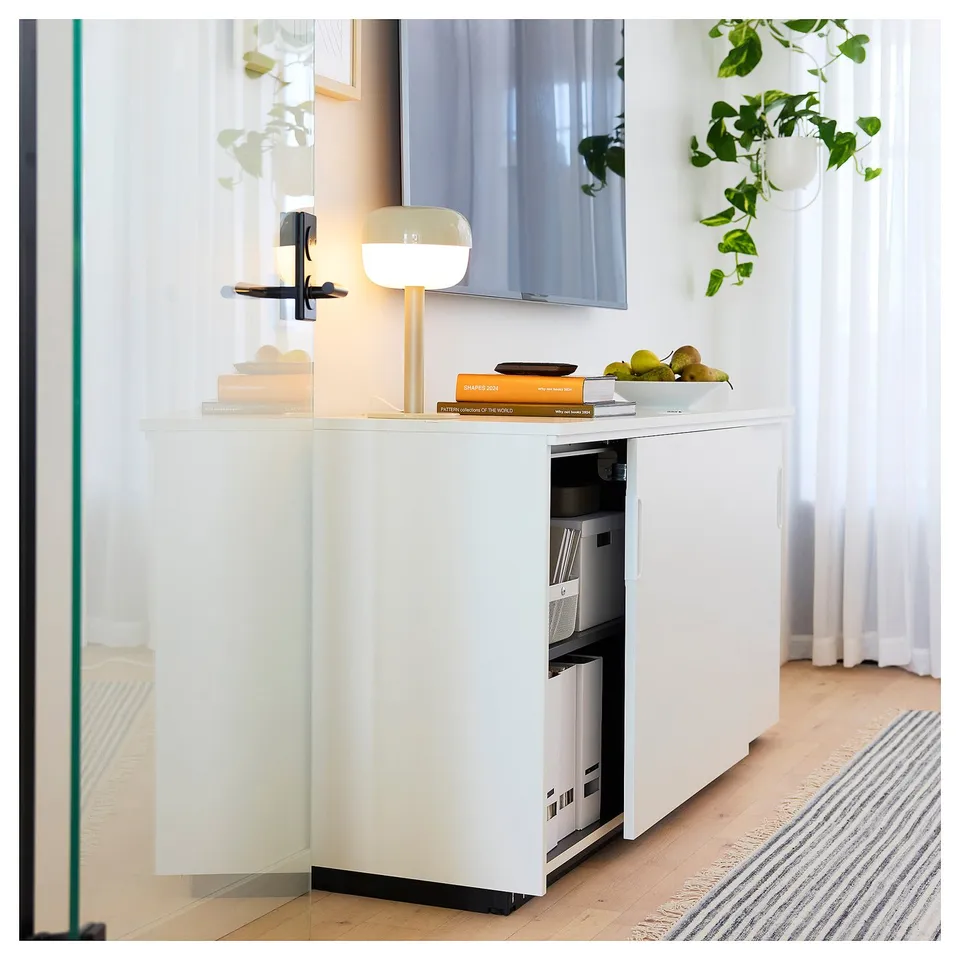 Modern white minimalist desk with drawers, featuring a lamp, books, and fruits on top. The desk has a clean design and adjustable shelves.