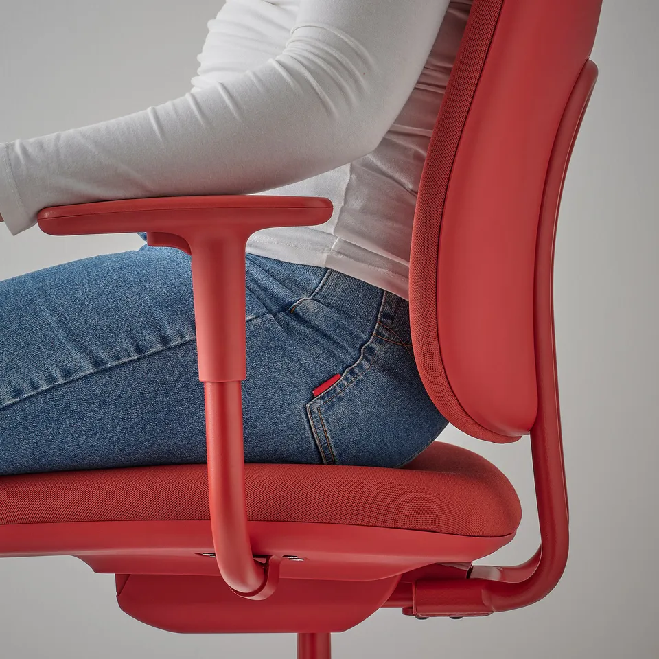 A person sits in a red office chair, adjusting the height of the armrests.