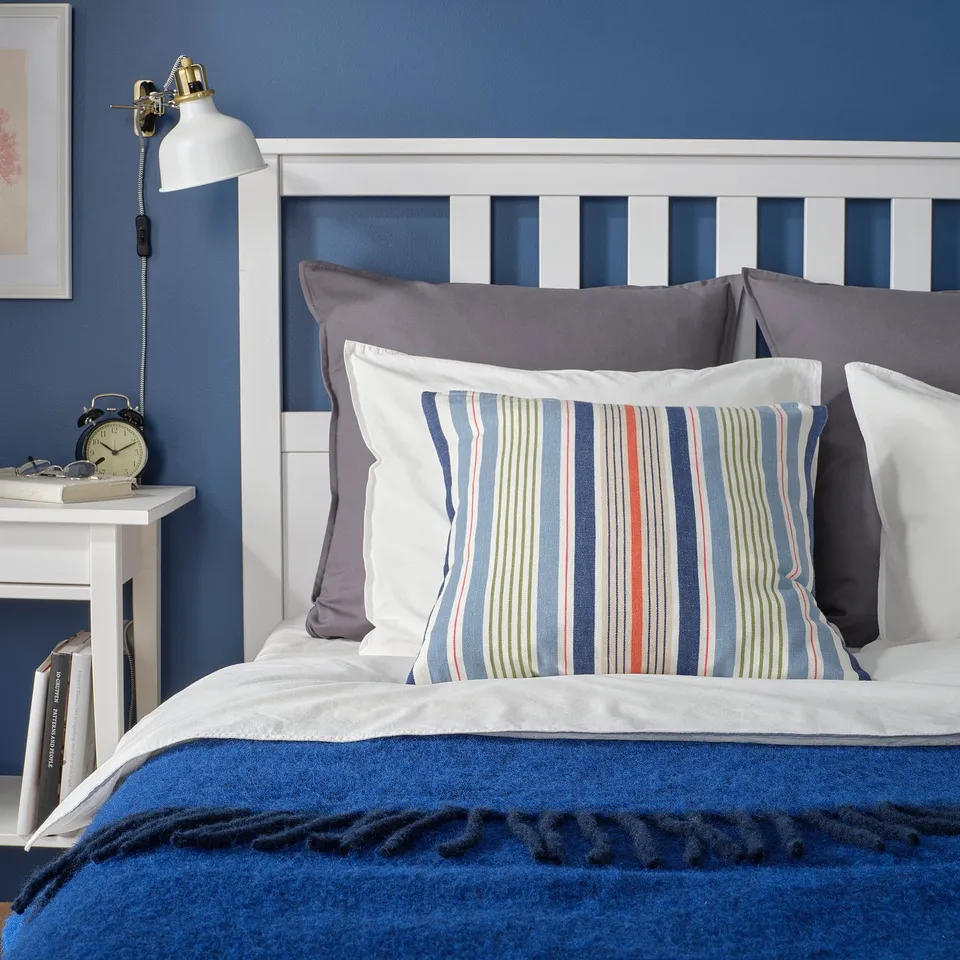 Blue bedroom with striped pillow, grey pillows, white bed frame, side table with lamp and alarm clock.