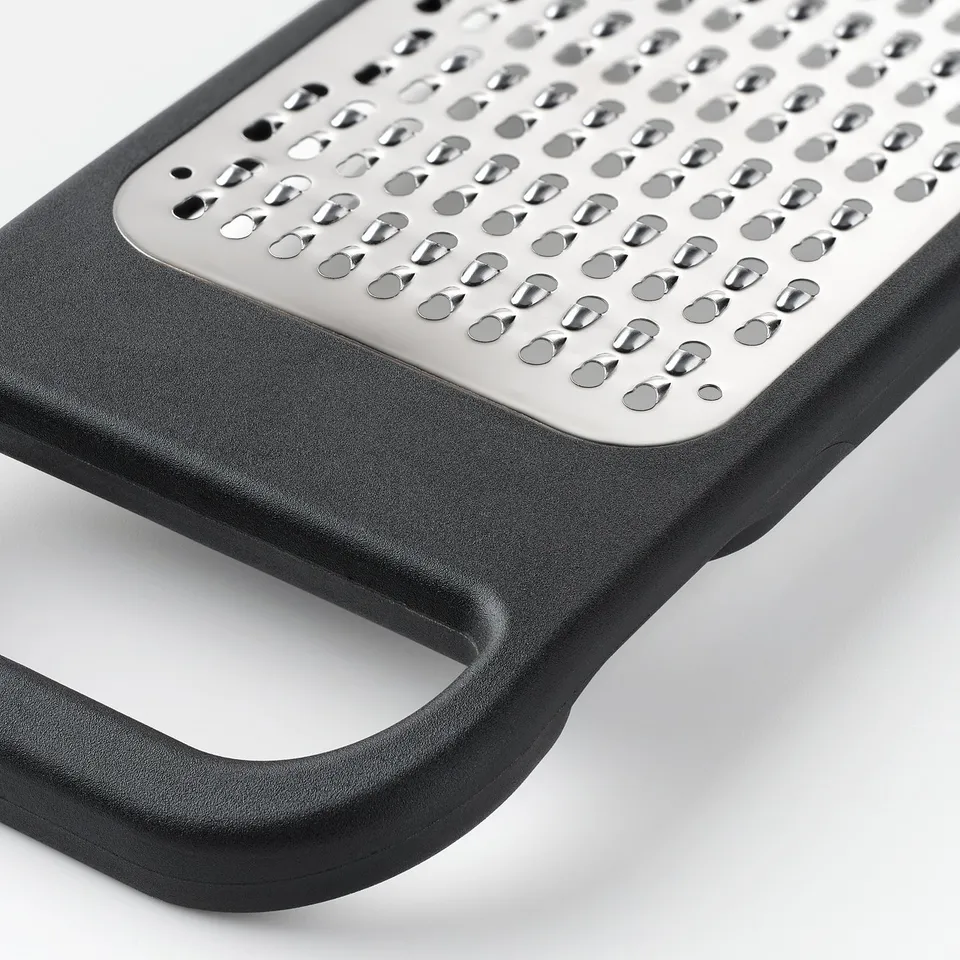Black plastic kitchen grater with sharp stainless steel holes, firm handle, placed on white surface.