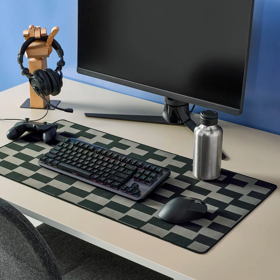 BLÅSKATA mouse pad, grey-black chequered, covers desk extensively, holds keyboard, mouse, and bottle, with monitor and gaming accessories on top.