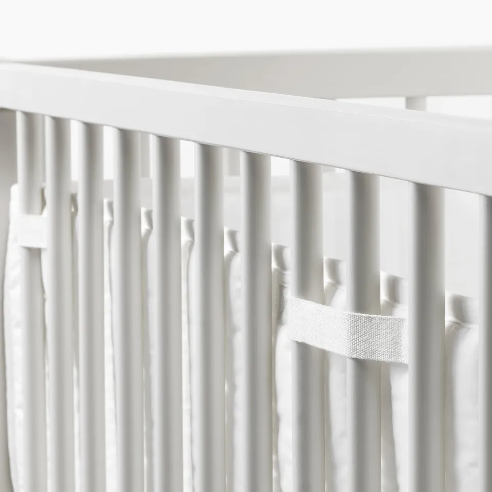 White crib bumper pad with loop fasteners, securing soft padding to prevent babys head, arms, or legs from hitting the cots hard spindles.