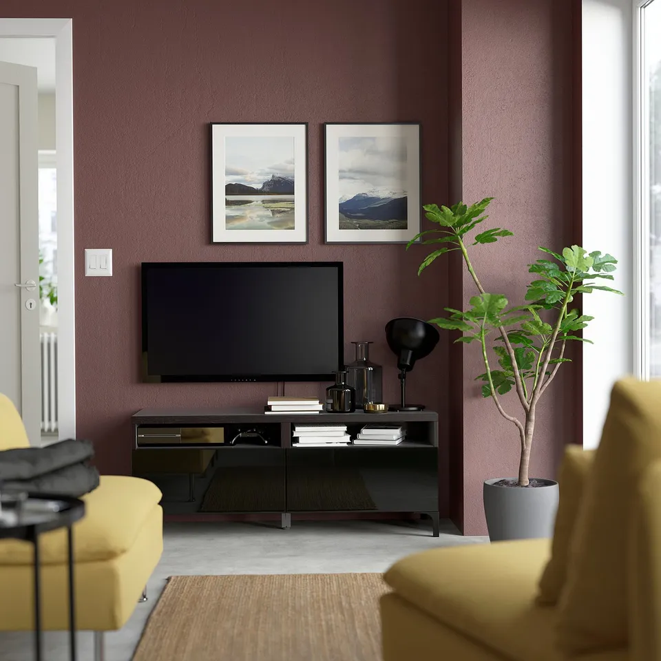 Living room with maroon walls, yellow sofa, flat screen tv on black stand, two framed landscape pictures, tall green plant.