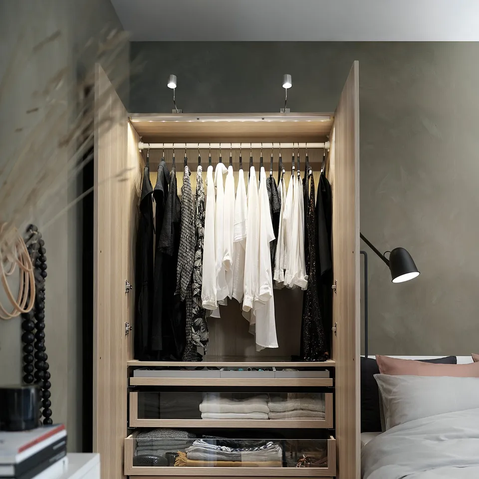 Open wooden closet with hangers, clothes, shelves, and drawers, lit from top, next to bed.