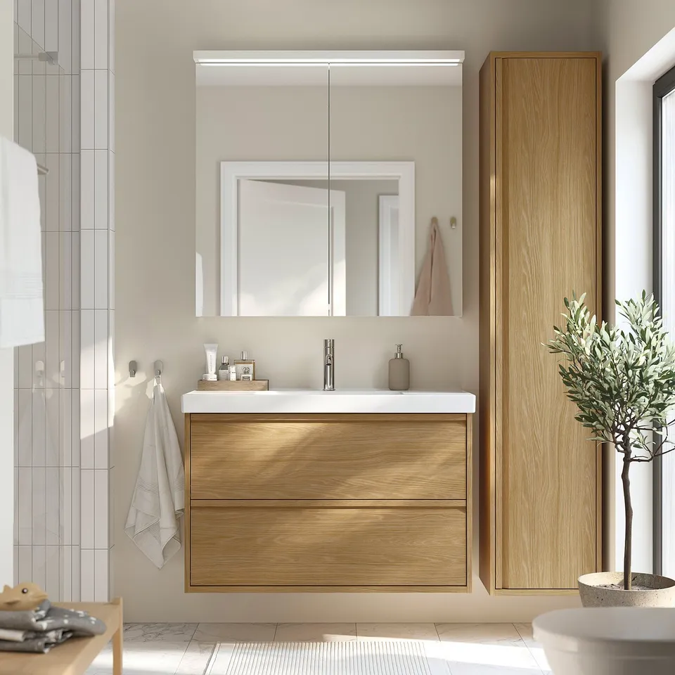 Modern bathroom with oak-finished ÄNGSJÖN vanity, minimal design, two drawers, sleek sink, backlit mirror, grey tiles, towel, plant.