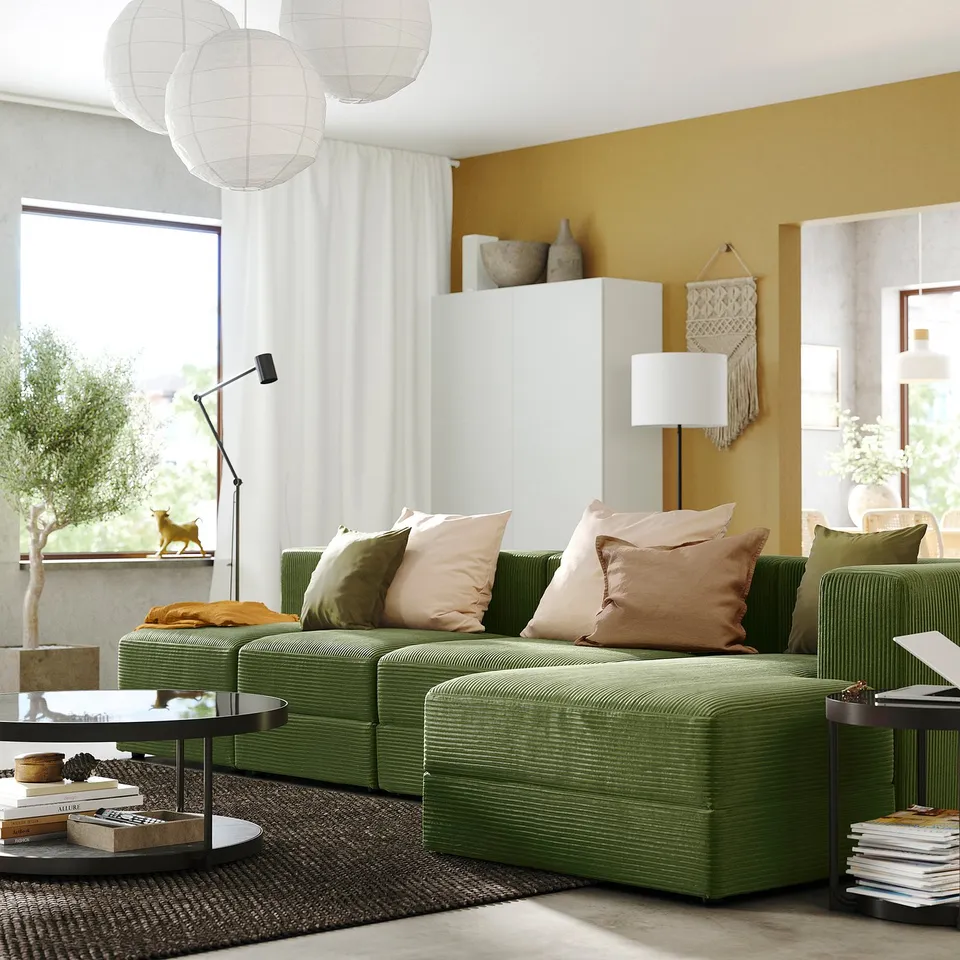 Modern living room: green JÄTTEBO sofa, pillows, floor lamp, books, cat on ledge, yellow wall.