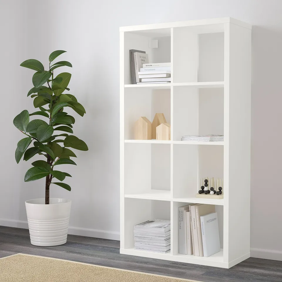 A white shelving unit, standing vertically, holding books and decorative items on its shelves. A potted plant is placed nearby on the floor.