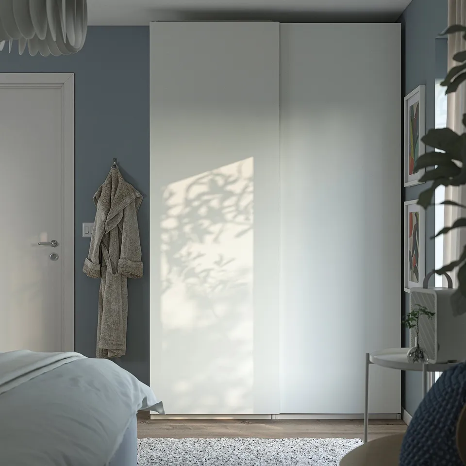 Modern bedroom with large white PAX wardrobe, hanging robe, potted plant, and bed with blue linens.