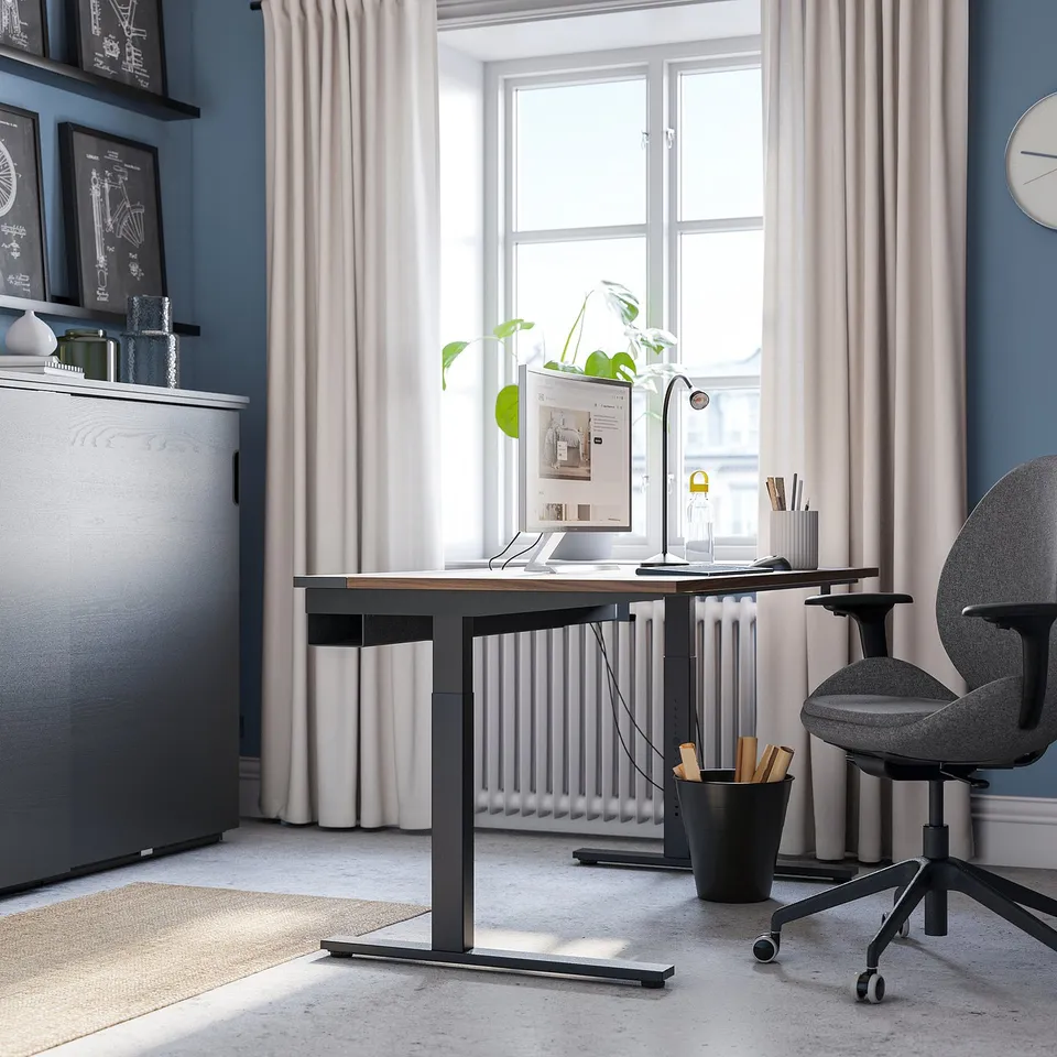 Modern home office setup with MITTZON adjustable desk in black and wood, grey chair, large window, white curtains, and wall shelf.