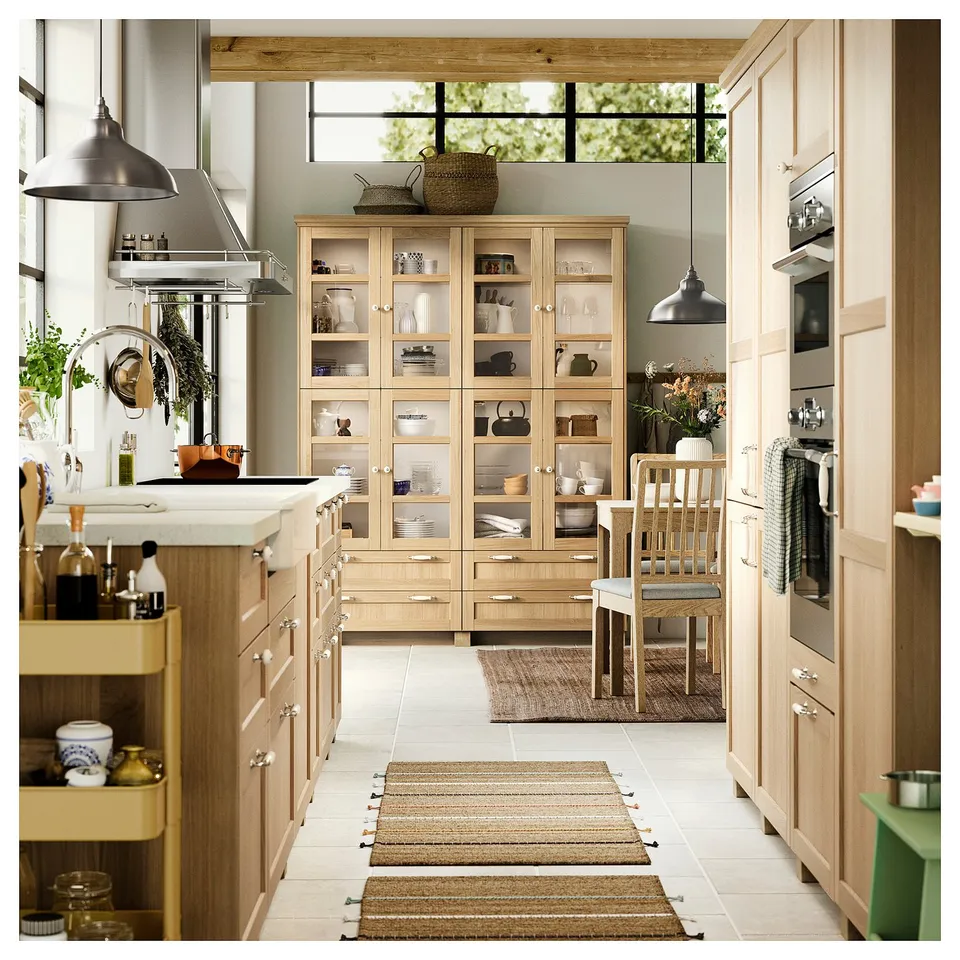 Modern kitchen with light wooden cabinets, central glass display unit, metallic fixtures, and decorative rugs.