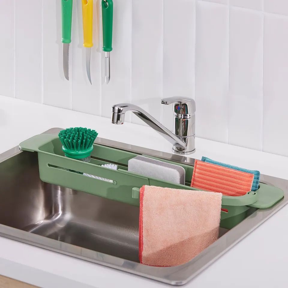 Stainless sink with green organiser holding scrubber, sponges, and towels, mounted on wall with hanging knives.