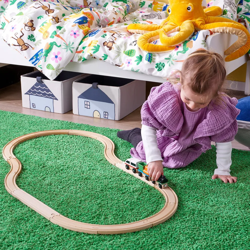 Child plays with wooden train set on green rug; locomotive and two wagons visible.