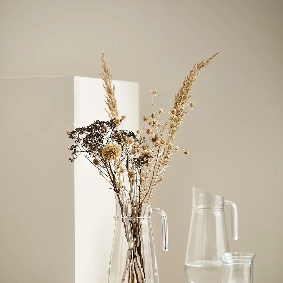 A clear glass vase holds dried flowers on a white stand. Beside it, glass pitchers feature simple, modern designs.