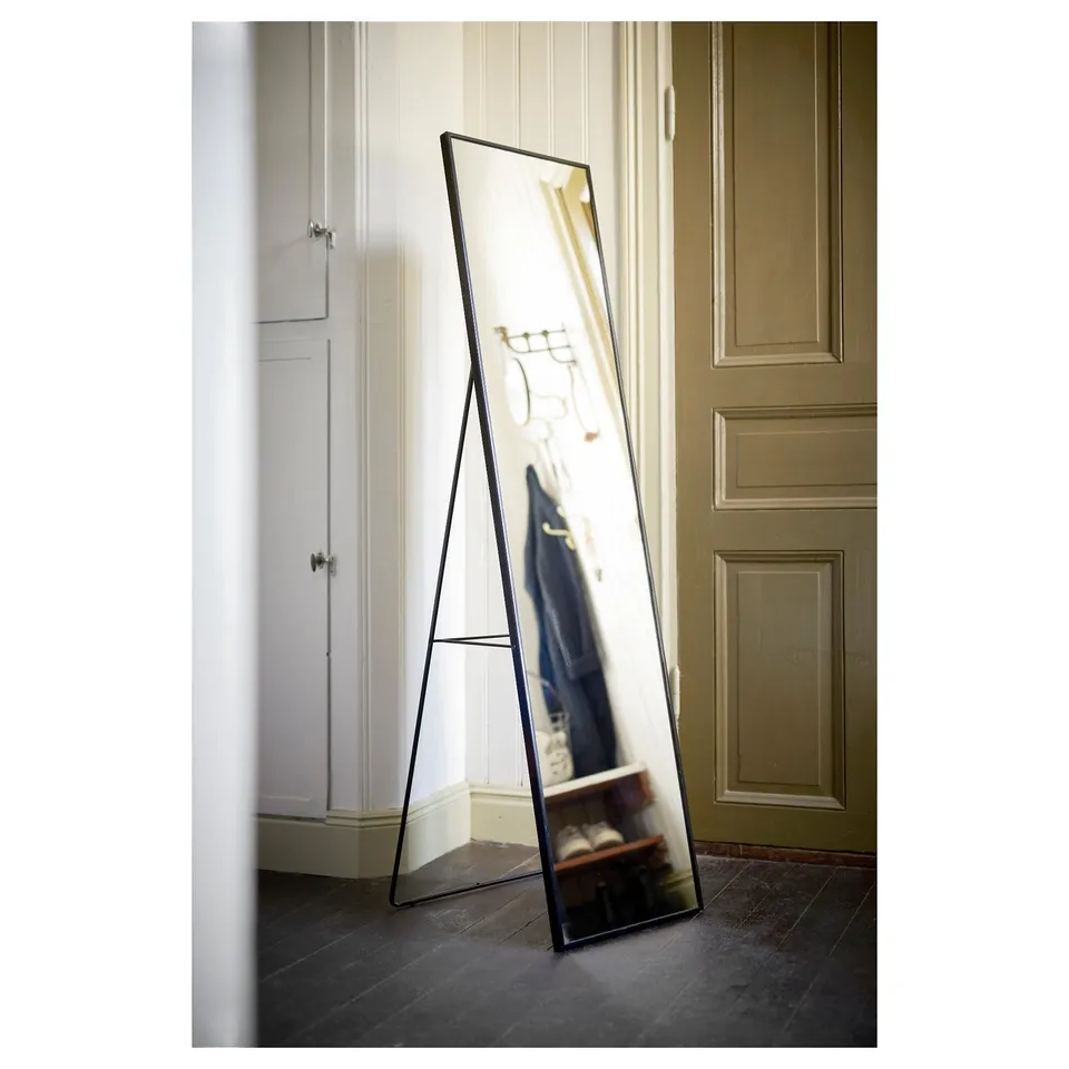 Black floor mirror with thin frame, leaning against wall, reflecting closet with clothes, in room with tiled floor and doors.