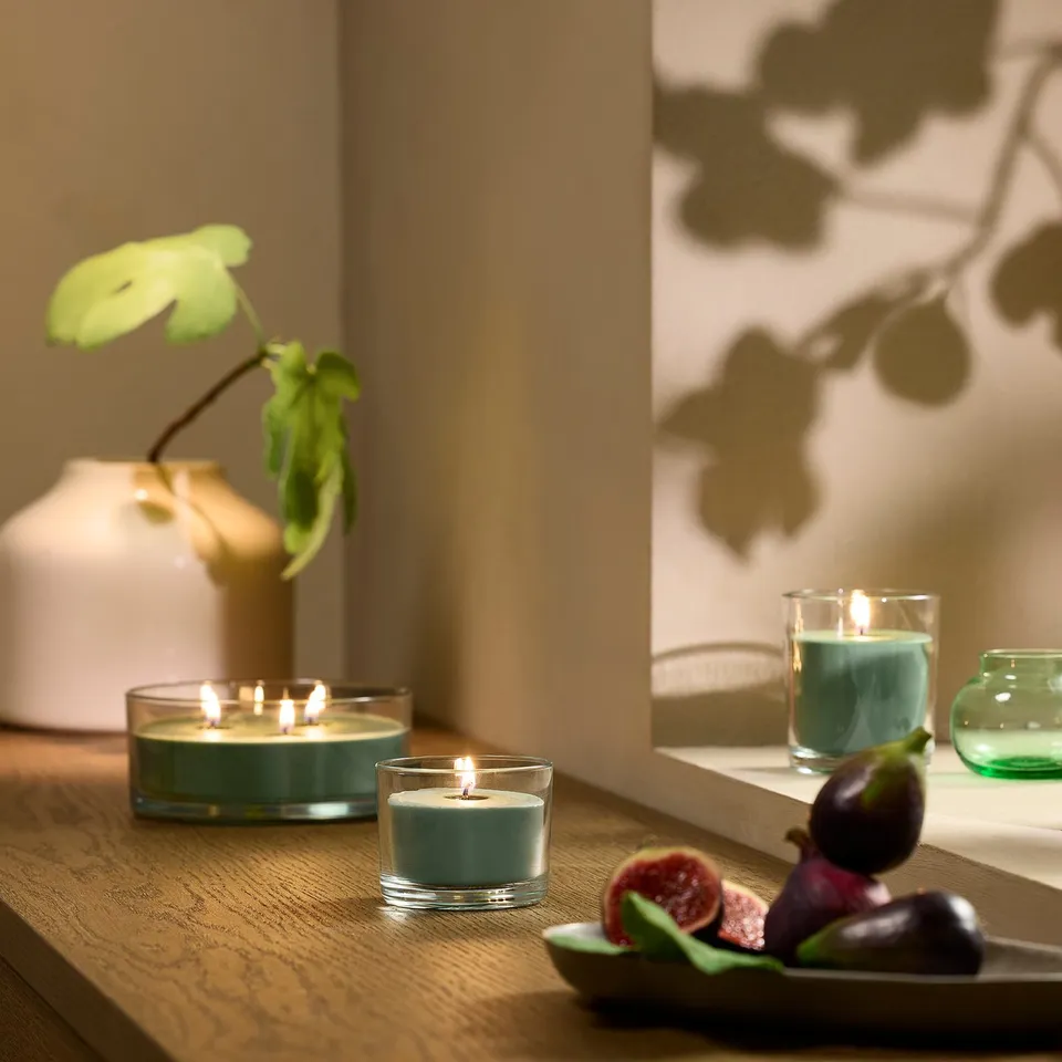 Cosy table setting with lit LOTSFÅGEL green candles, fresh figs, and white vase with bright green plant, creating a serene atmosphere.