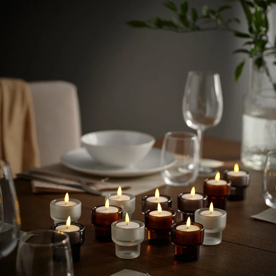 Elegant dinner setup with lit SKATGÅS led tealights, white & brown glass, surrounding plates, bowls, glasses, and greenery in a cosy setting.