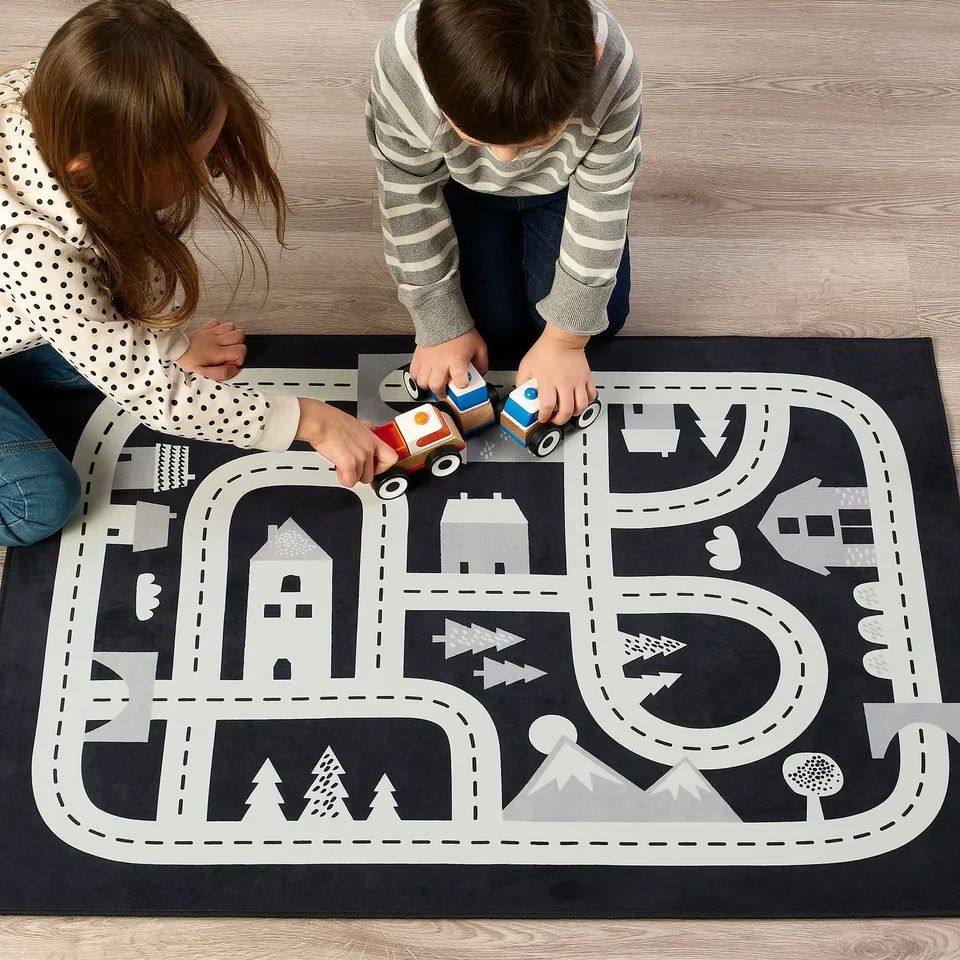Two kids playing with toy cars on a black and white road rug. The rug houses, trees, and winding roads.