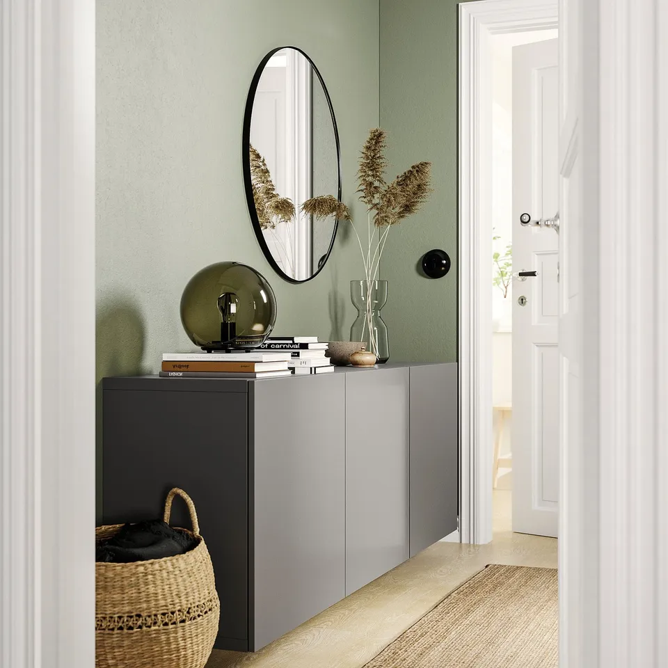 Modern entryway with BESTÅ grey storage, round black mirror, green lamp, woven basket, and dried flowers in vase.